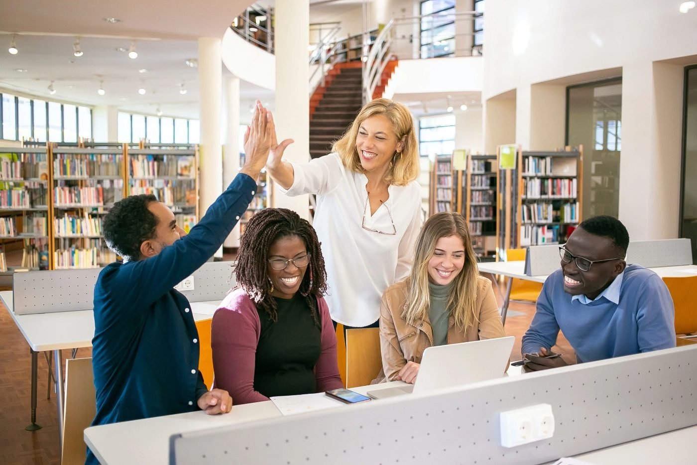 A teacher high-fiving a student while classmates smile around a table in a library